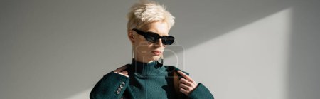 Téléchargez les photos : Modèle blond dans des lunettes de soleil à la mode portant un manteau vert sur fond gris, bannière - en image libre de droit
