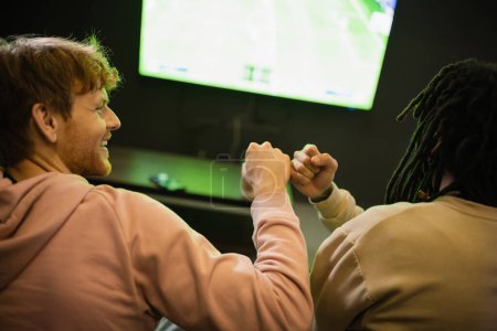 Smiling man doing fist bump with multiracial friend in gaming club 