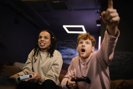 Photo for KYIV, UKRAINE - FEBRUARY 13, 2023: Excited man pointing with finger while playing video game with multiracial friend in gaming club - Royalty Free Image