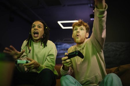 Photo for KYIV, UKRAINE - FEBRUARY 13, 2023: Low angle view of excited multiracial man playing video game with friend in gaming club - Royalty Free Image