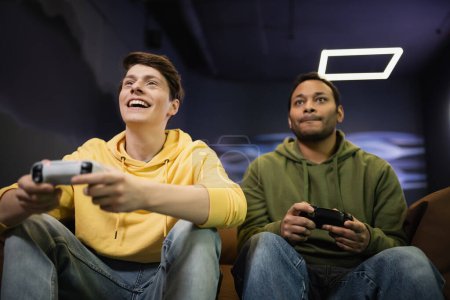 Photo for KYIV, UKRAINE - FEBRUARY 13, 2023: Low angle view of cheerful man playing video game with indian friend in gaming club - Royalty Free Image