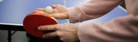 Cropped view of man holding ball and racket while playing table tennis in gaming club, banner 
