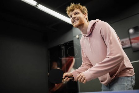 Téléchargez les photos : Vue à angle bas de l'homme rousse souriant jouant au tennis de table dans un club de jeu - en image libre de droit