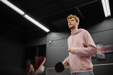 Téléchargez les photos : Jeune homme rousse avec raquette jouant au tennis de table dans un club de jeux - en image libre de droit