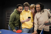 Multiethnic friends smiling while using smartphone near table tennis in gaming club  Poster #650688462