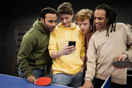 Cheerful multiethnic friends using cellphone near table tennis in gaming club  Poster 650688472