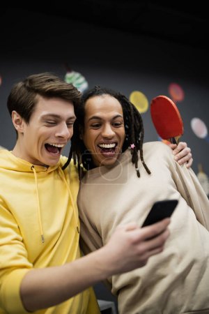 Amis interraciaux positifs avec raquette de tennis en utilisant le téléphone mobile dans le club de jeu 