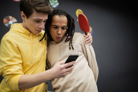 Choqué amis multiethniques avec raquette de tennis en utilisant smartphone dans le club de jeu 