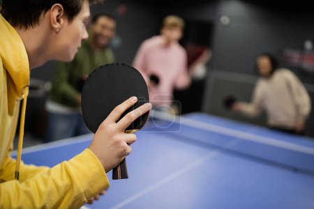 Young man playing table tennis with blurred friends in gaming club 