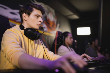 Young man with headphones playing video game on computer near blurred friends in cyber club 