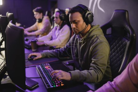 Photo for Indian gamer in headphones with microphone playing video game on computer near blurred friends in gaming club - Royalty Free Image