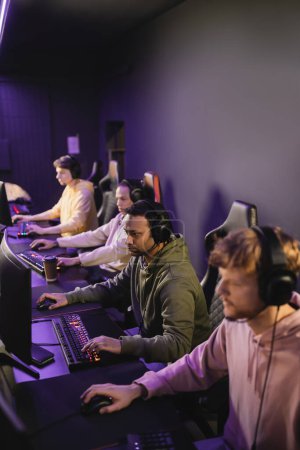 Photo for Indian gamer in headphones with microphone playing video game on computer near team in cyber club - Royalty Free Image
