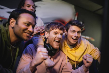 Podekscytowany mężczyzna ze słuchawkami odwracając wzrok w pobliżu międzyrasowych przyjaciół w klubie gier 