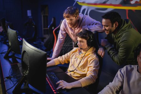 Téléchargez les photos : Amis multiethniques debout près de gamer dans les écouteurs jouer à un jeu vidéo sur ordinateur dans le cyber-club - en image libre de droit