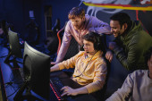 Multiethnic friends standing near gamer in headphones playing video game on computer in cyber club  t-shirt #650689688