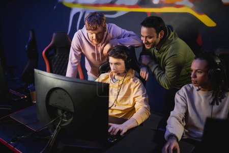 Téléchargez les photos : Des hommes souriants et multiethniques regardant un ami jouer à un jeu vidéo dans un club de jeu - en image libre de droit