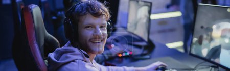 Cheerful young gamer in headphones looking at camera near computer in gaming club, banner 
