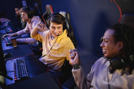 Cheerful man in headphones doing fist bump with multiracial friend in gaming club 