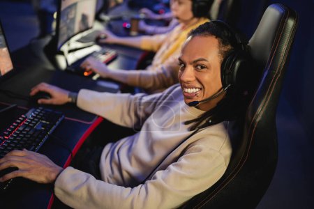 Smiling multiracial gamer in headphones looking at camera while playing video game with team in gaming club 