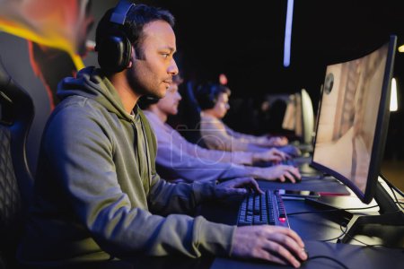 Photo for Side view of indian gamer in headphones playing video game with blurred team in club - Royalty Free Image