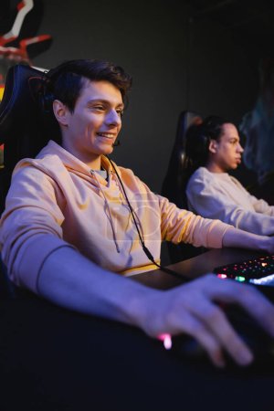 Smiling player with headphones sitting near keyboard in cyber club 