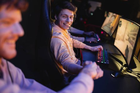 Hombre sonriente en auriculares estrechando la mano de un amigo borroso cerca de las computadoras en el club cibernético 