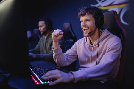 Foto de Emocionado joven jugador en los auriculares que muestran sí gesto cerca de la computadora en el club de juegos - Imagen libre de derechos