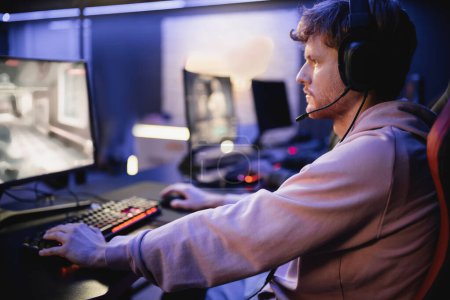 Téléchargez les photos : Vue latérale du jeune homme dans les écouteurs avec microphone jouer au jeu vidéo dans le cyber-club - en image libre de droit