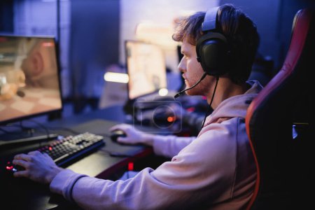 Téléchargez les photos : Vue latérale du jeune joueur dans les écouteurs jouer au jeu d'ordinateur dans le cyber-club - en image libre de droit