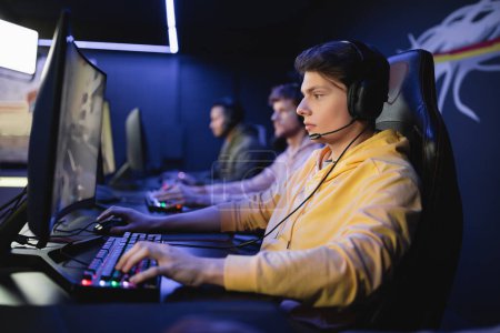 Téléchargez les photos : Vue latérale du joueur dans les écouteurs jouant au jeu vidéo sur ordinateur avec l'équipe dans le cyber-club - en image libre de droit