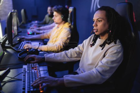 Foto de Jugador multirracial jugando videojuego en la computadora cerca del equipo borroso en el club cibernético - Imagen libre de derechos