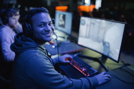 Foto de Sonriente jugador indio en auriculares mirando a la cámara cerca de la computadora en el club cibernético con iluminación - Imagen libre de derechos