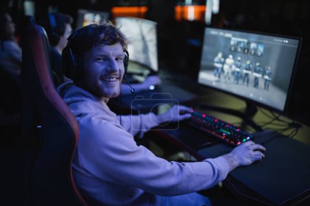 Joueur insouciant dans un casque jouant à un jeu vidéo sur ordinateur dans un cyber-club avec éclairage 