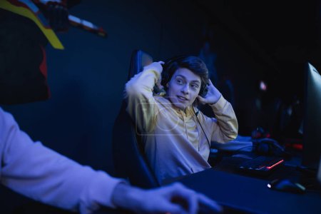 Junger Gamer mit Kopfhörer spricht mit verschwommenem Freund in Cyber-Club mit Beleuchtung 
