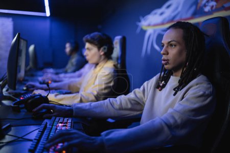 Téléchargez les photos : Joueur multiracial jouant à un jeu vidéo avec des amis flous dans le cyber-club - en image libre de droit
