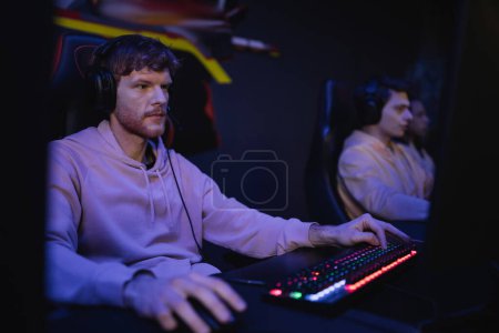 Gamer mit Kopfhörern spielt Videospiel mit verschwommenen Freunden im Cyber-Club 