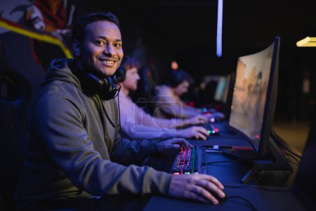 Photo for Smiling indian gamer looking at camera near computer in cyber club - Royalty Free Image