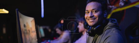 Photo for Positive indian gamer with headphones looking at camera in cyber club, banner - Royalty Free Image