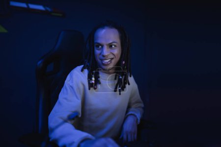 Lächelnde multirassische Spieler mit Dreadlocks sitzen in Cyber-Club mit Beleuchtung 