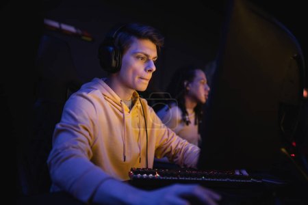 Hombre joven en auriculares jugando juego de ordenador en el club cibernético con iluminación 