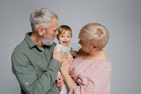 Photo for Carefree toddler girl looking at camera near bearded grandfather and granny isolated on grey - Royalty Free Image