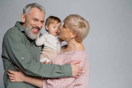 mature woman kissing granddaughter near pleased bearded husband isolated on grey