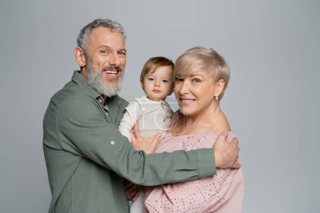 Téléchargez les photos : Grands-parents heureux regardant la caméra près de petite-fille bien-aimée isolé sur gris - en image libre de droit