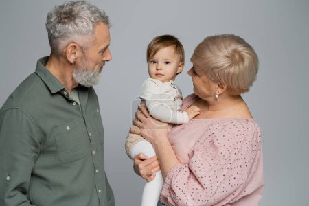 senior woman holding toddler granddaughter near bearded grey haired husband isolated on grey