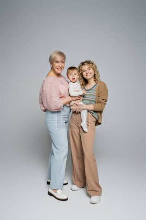 Téléchargez les photos : Pleine longueur de femme mature souriant à la caméra près de fille adulte avec tout-petit enfant sur fond gris - en image libre de droit