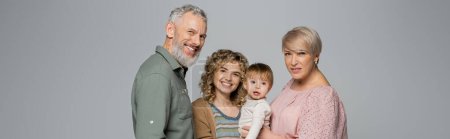 joyful middle aged couple smiling near adult daughter with toddler girl isolated on grey, banner