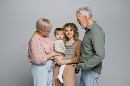 senior couple looking at cheerful adult daughter with toddler child isolated on grey