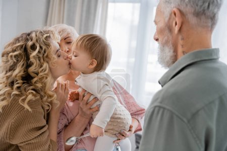 blonde woman kissing beloved daughter near mature parents at home
