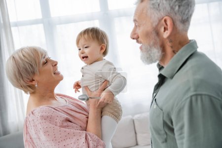 happy mature woman holding beloved granddaughter near bearded husband at home