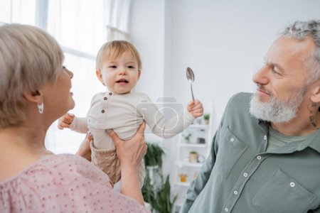 woman holding cheerful granddaughter with spoon near bearded grey haired husband in kitchen